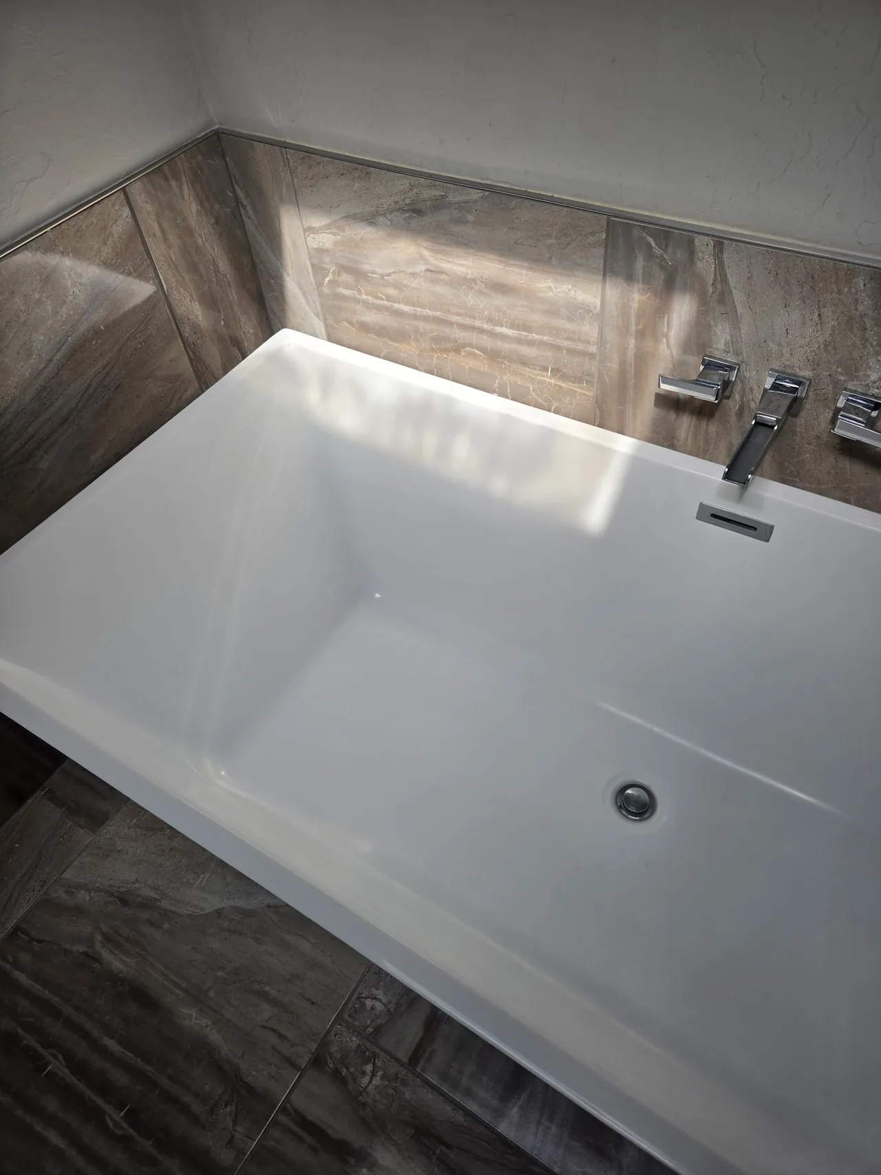 White soaking tub with chrome fixtures and stone patterned tile surround